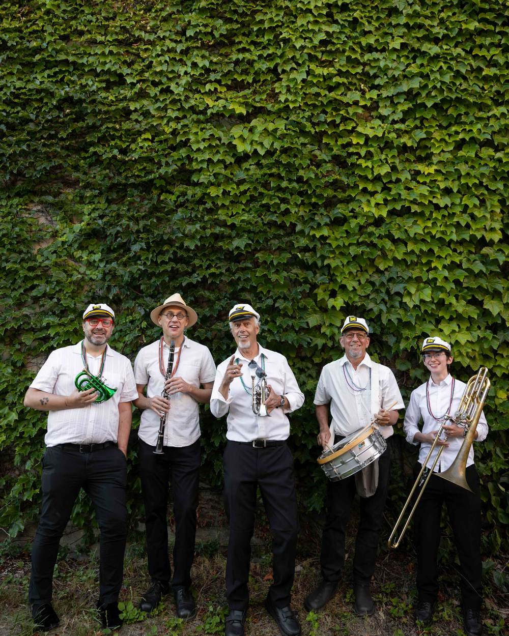 The OompahME quintet in white shirts and captain’s hats with their instruments, in front of an ivy wall