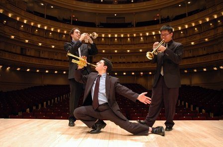 Three trumpet players in dark suits posing on the stage of a concert hall