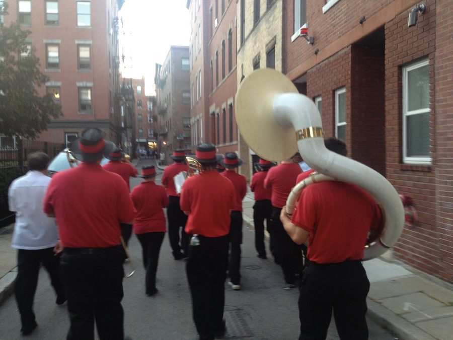 A marching band in red shirts and black hats walking down a city street, seen from behind
